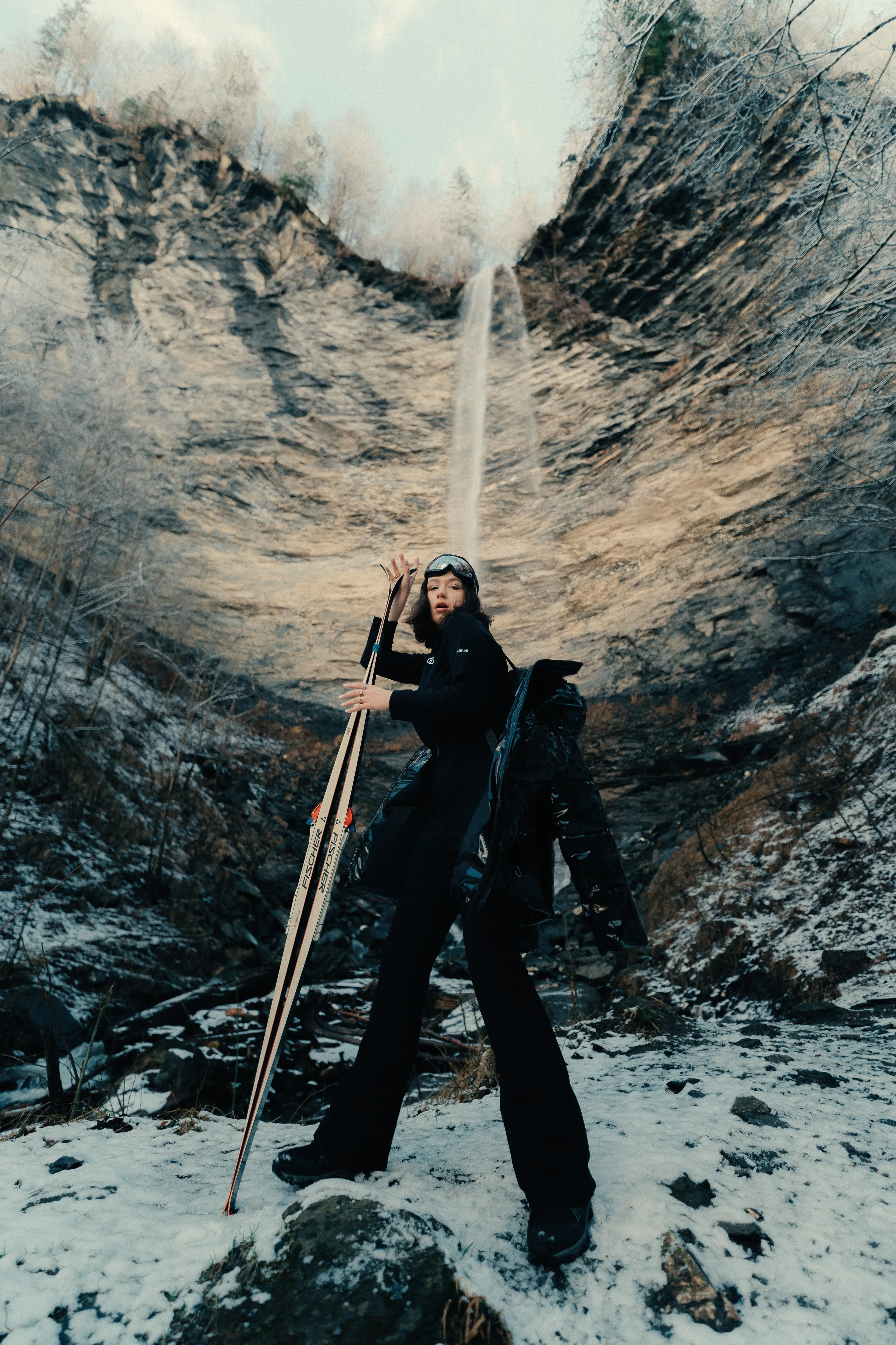 Une modèle en combinaison noire pose avec un bâton de ski devant une cascade gelée, au pied d'une falaise.
