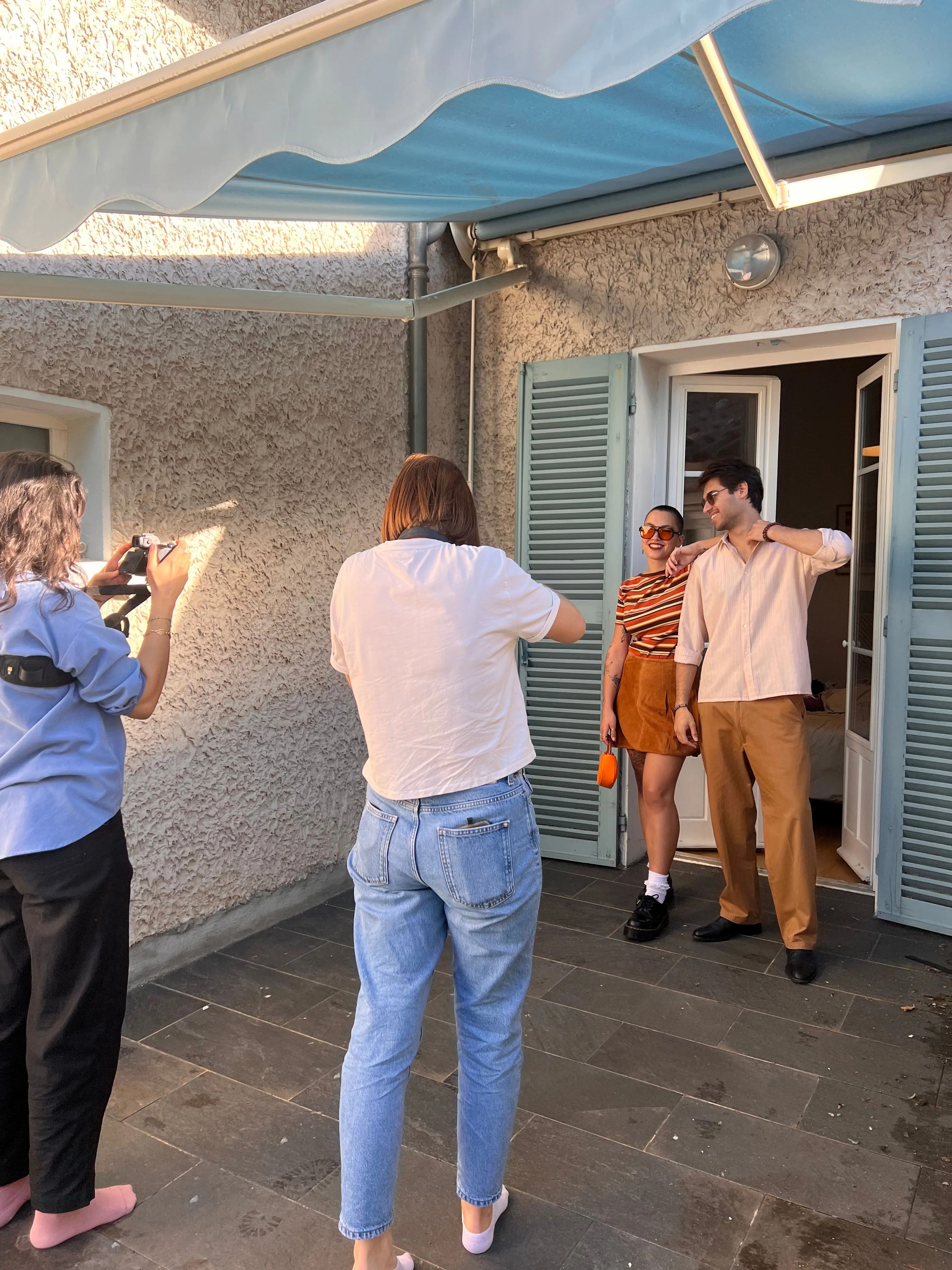 Des photographes shootent un duo de modèles devant une porte-fenêtre aux volets bleus.