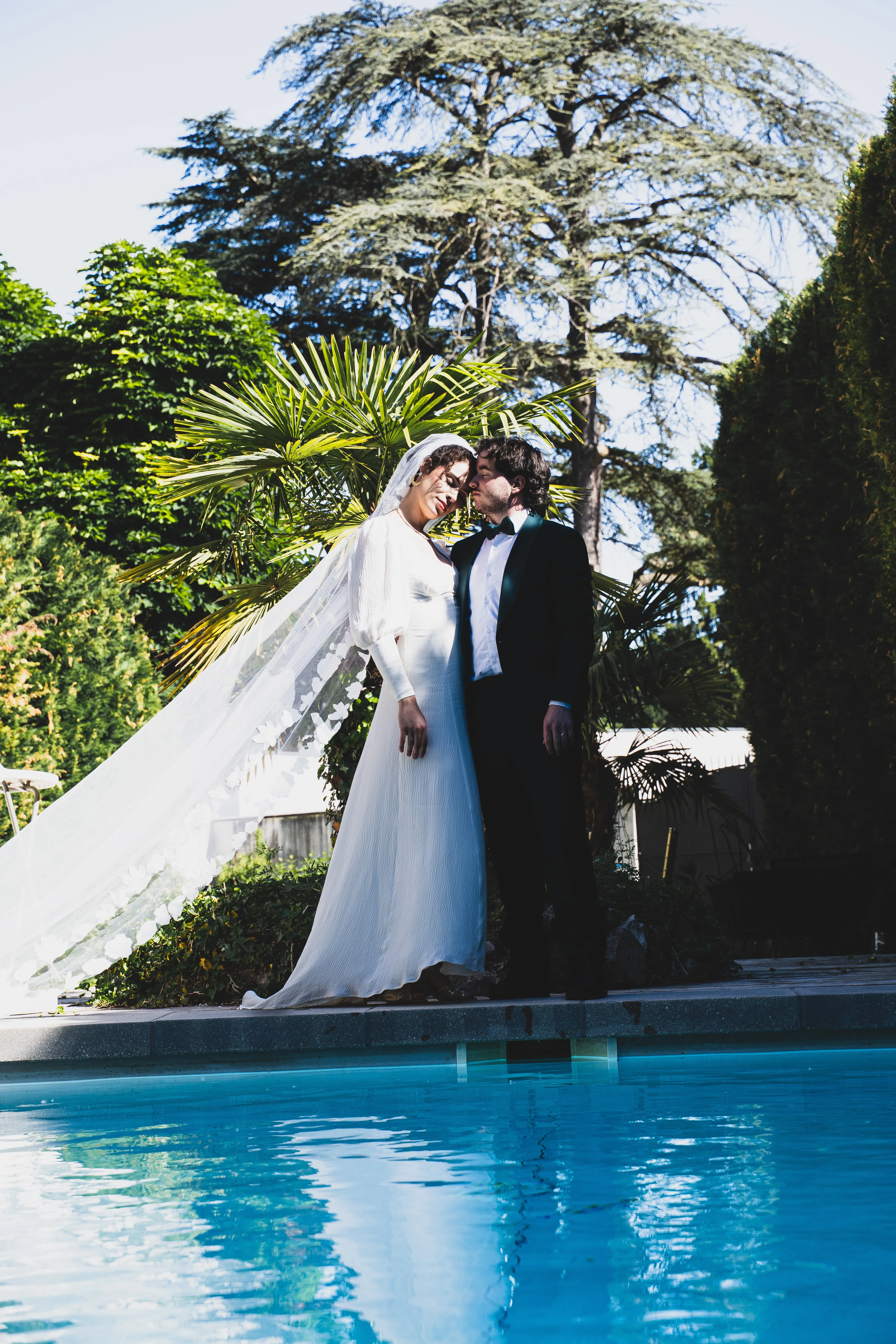 Les mariés enlacés au bord de la piscine du manoir, voile au vent, sous un grand cèdre.