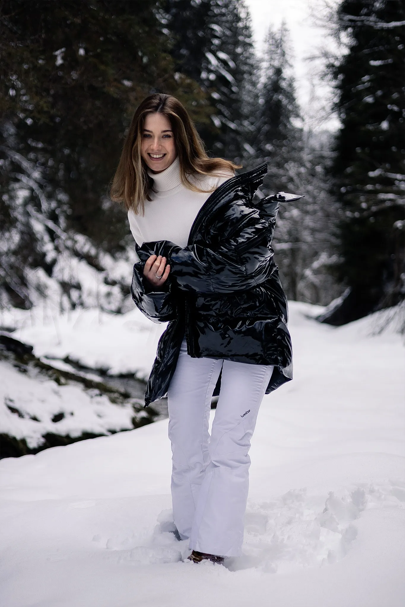 Une modèle en pantalon de ski blanc serre une doudoune noire brillante, dans une forêt enneigée.