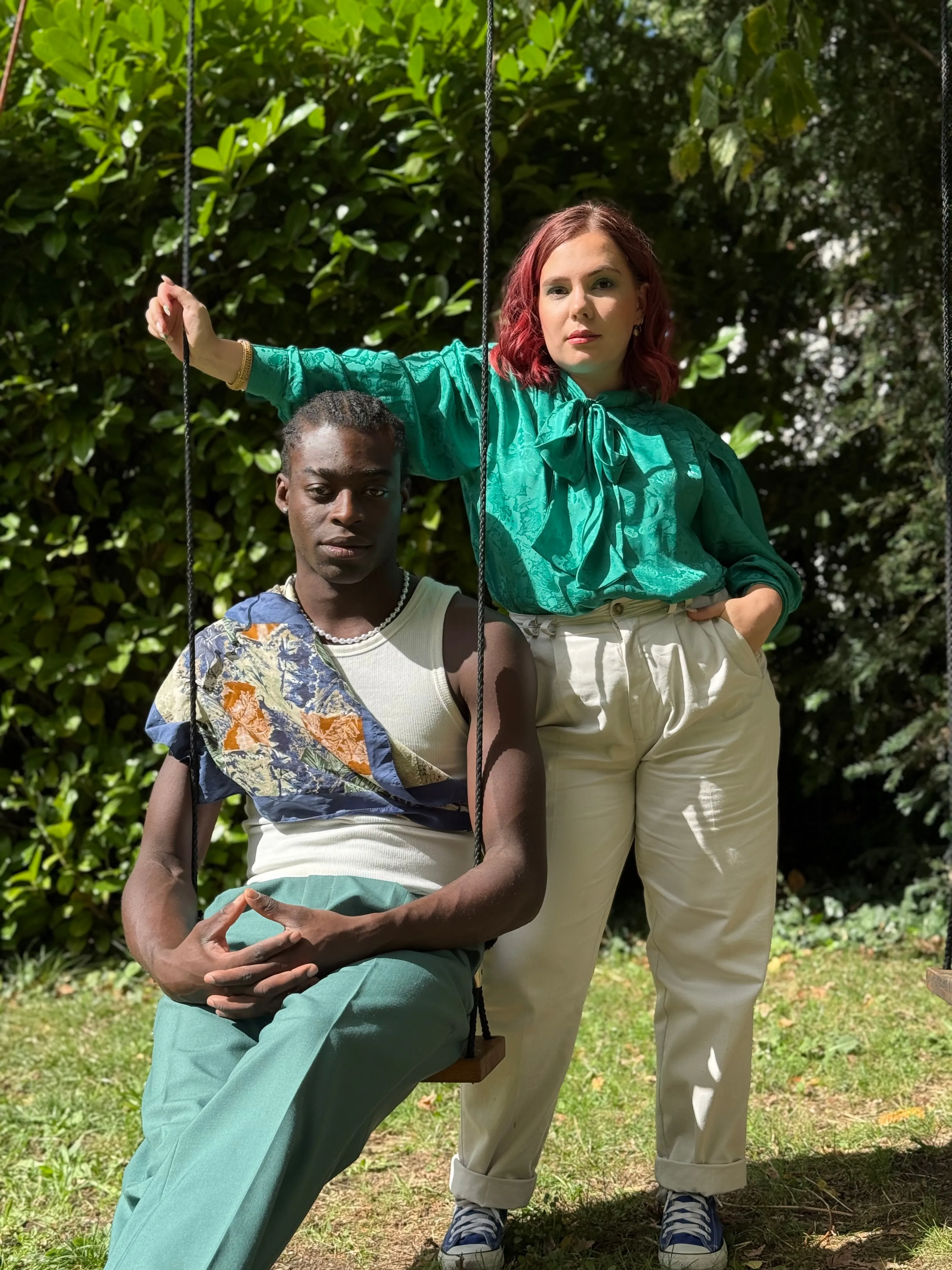 Un modèle en chemise à motifs assis sur une balançoire et une modèle en chemisier vert, dans un jardin.