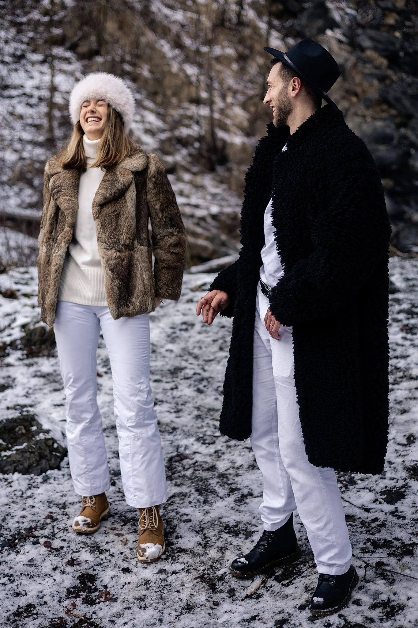Une modèle en veste de fourrure et bonnet blanc rit à côté d'un modèle en long manteau noir, dans un sous-bois enneigé.