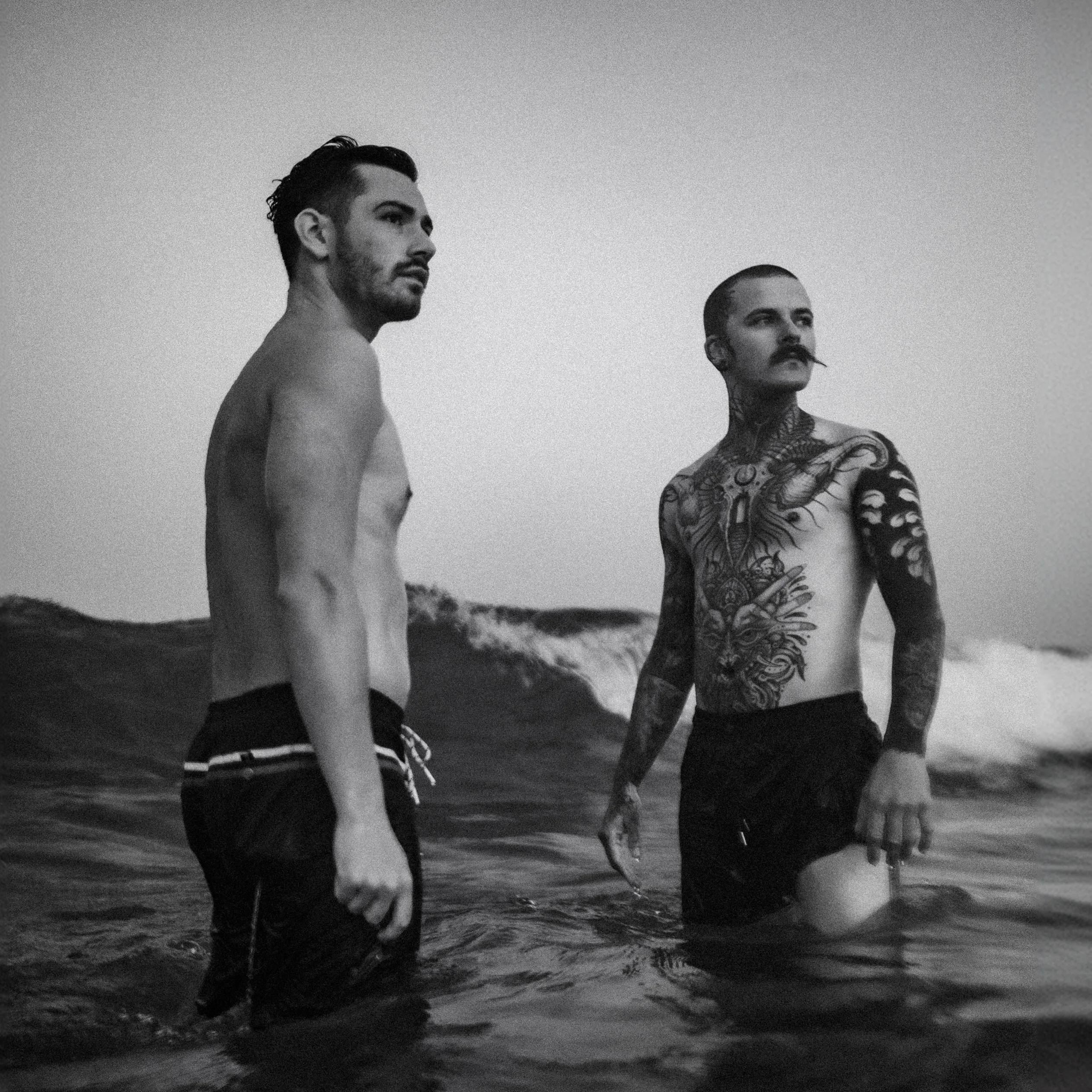 Deux modèles torse nu debout dans la mer, face aux vagues, en noir et blanc.