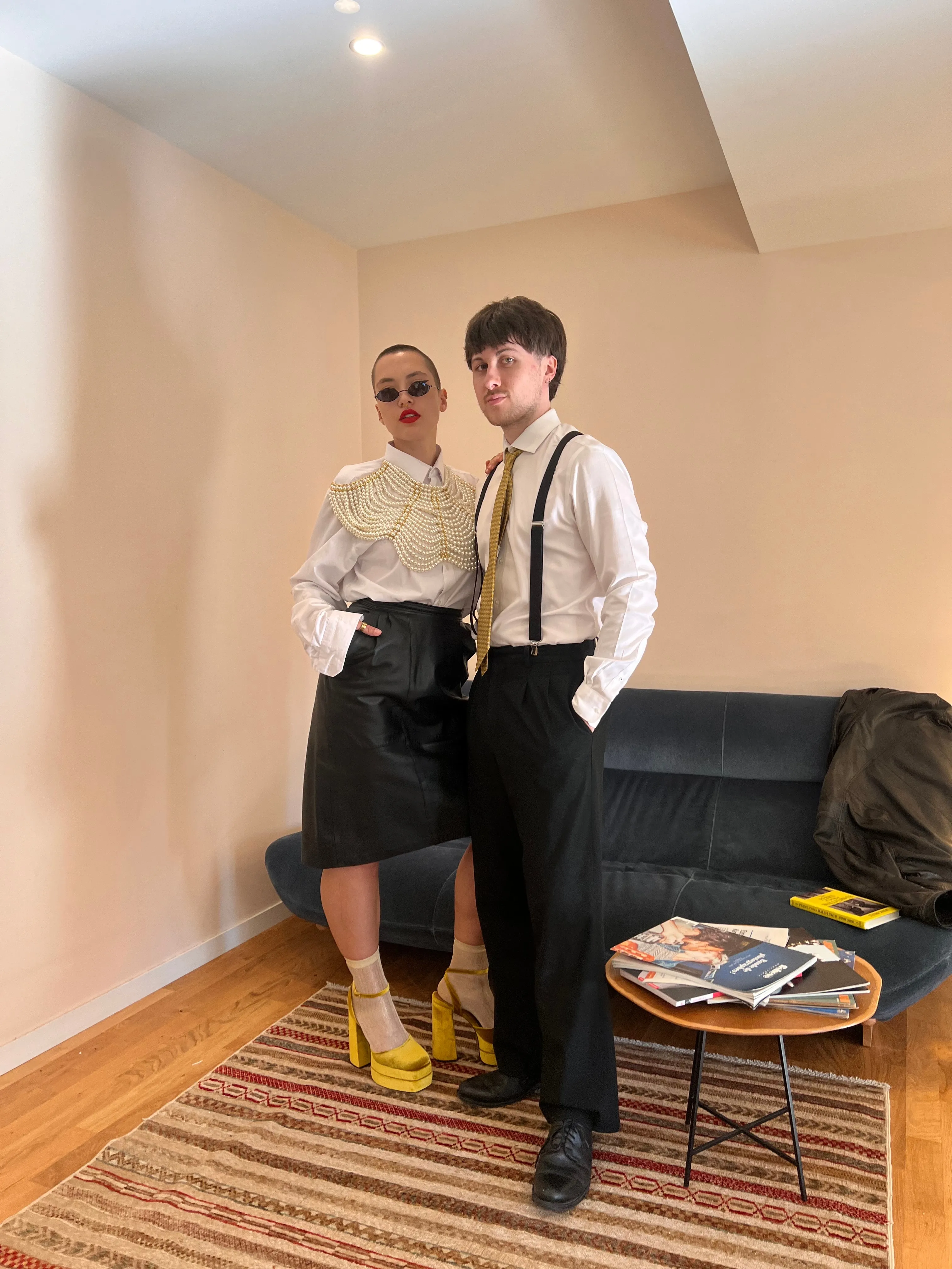 Un duo en chemises blanches, jupe en cuir et bretelles, pose dans un salon aux tapis rayés.