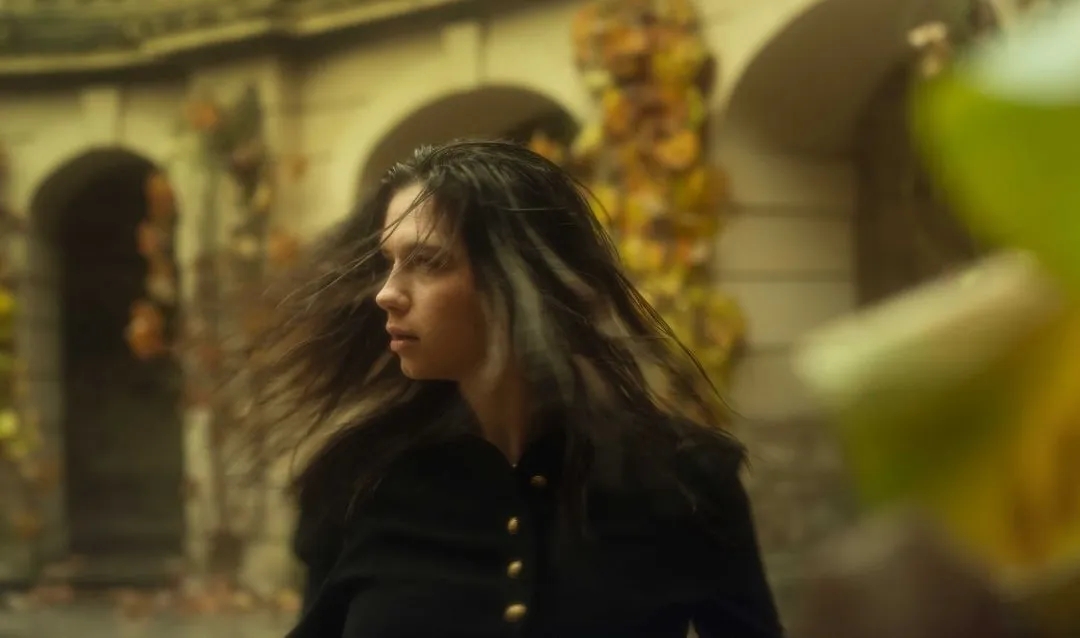 Une modèle en manteau noir, cheveux en mouvement, devant des arcades couvertes de feuillage d'automne à Prague.