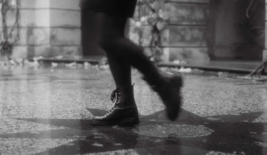 Une modèle en bottines traverse une cour pavée et mouillée, en noir et blanc, à Prague.