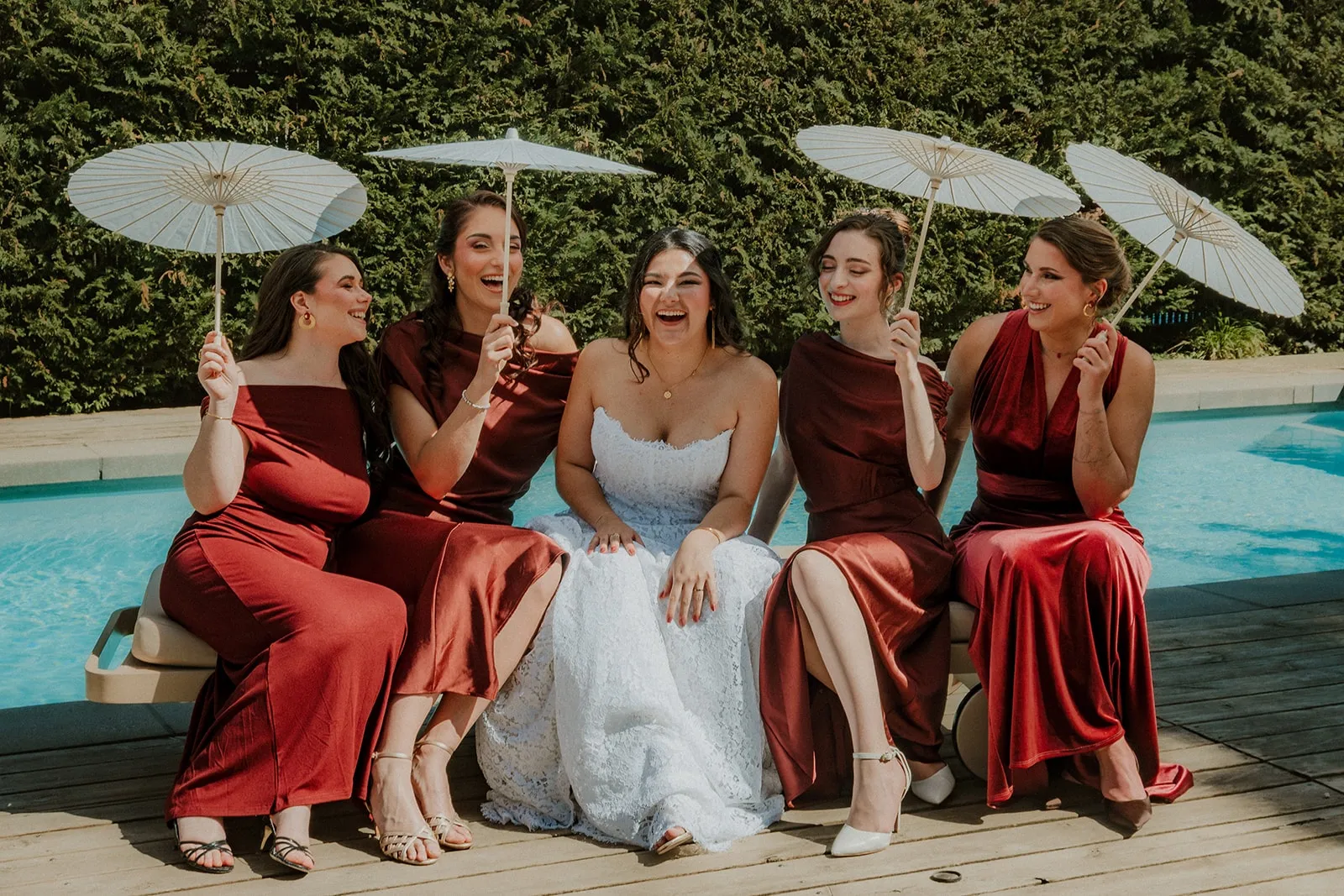 La mariée en dentelle et ses demoiselles d'honneur en robes bordeaux, ombrelles à la main, riant au bord de la piscine.