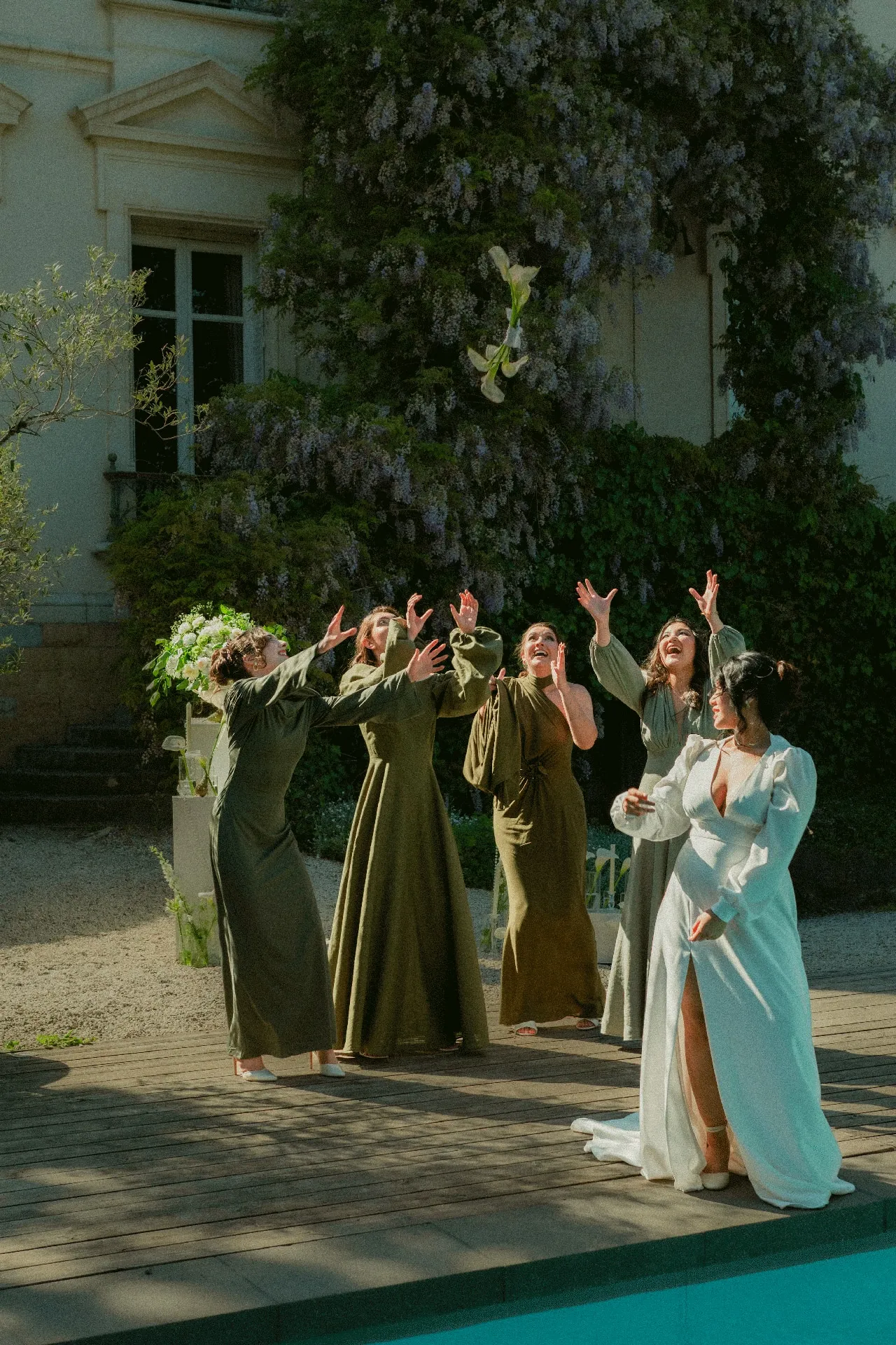 La mariée lance son bouquet d'arums vers ses demoiselles d'honneur en robes vert olive, devant la façade couverte de glycine.