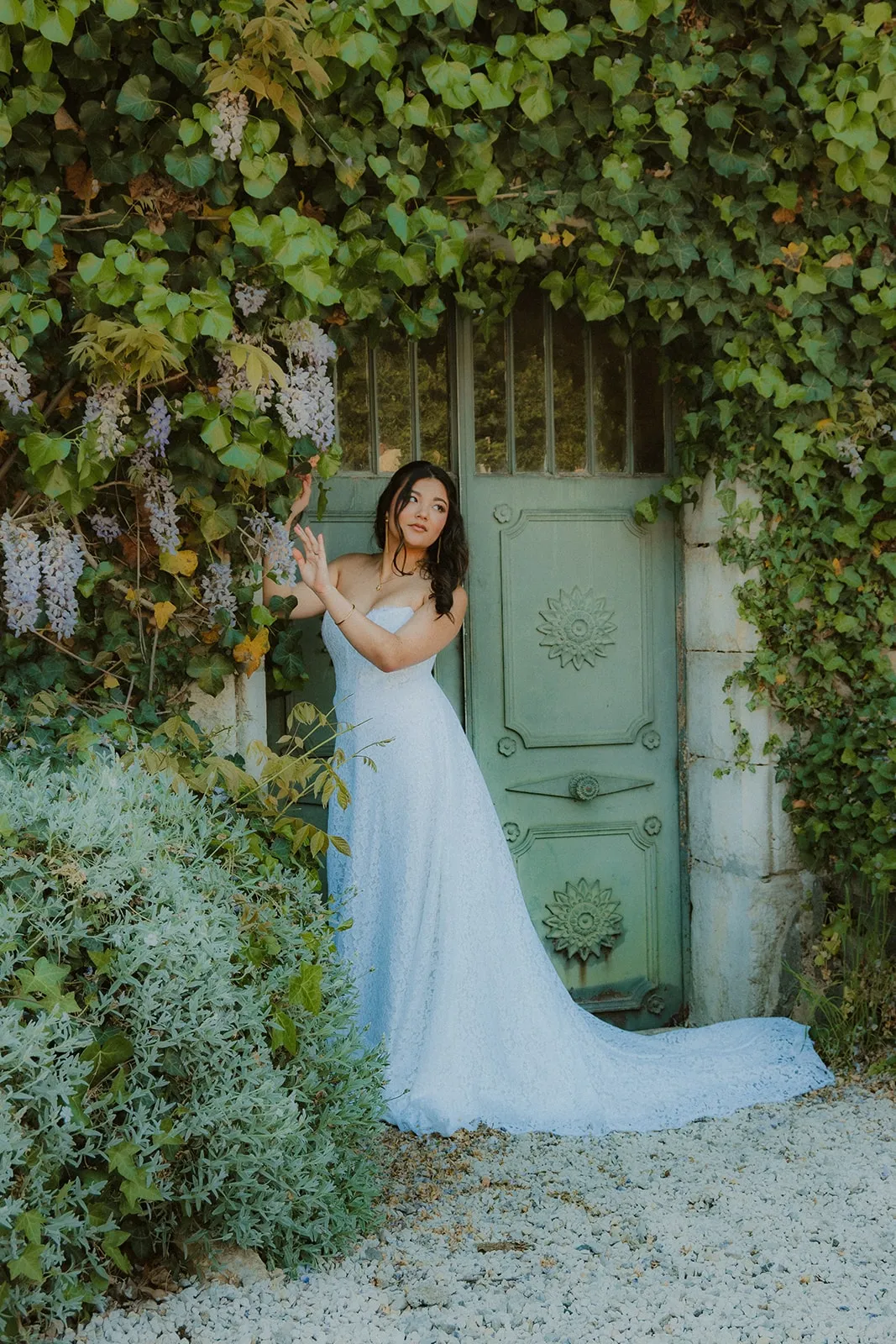 Une mariée en robe longue pose devant une porte verte encadrée de glycine et de lierre.