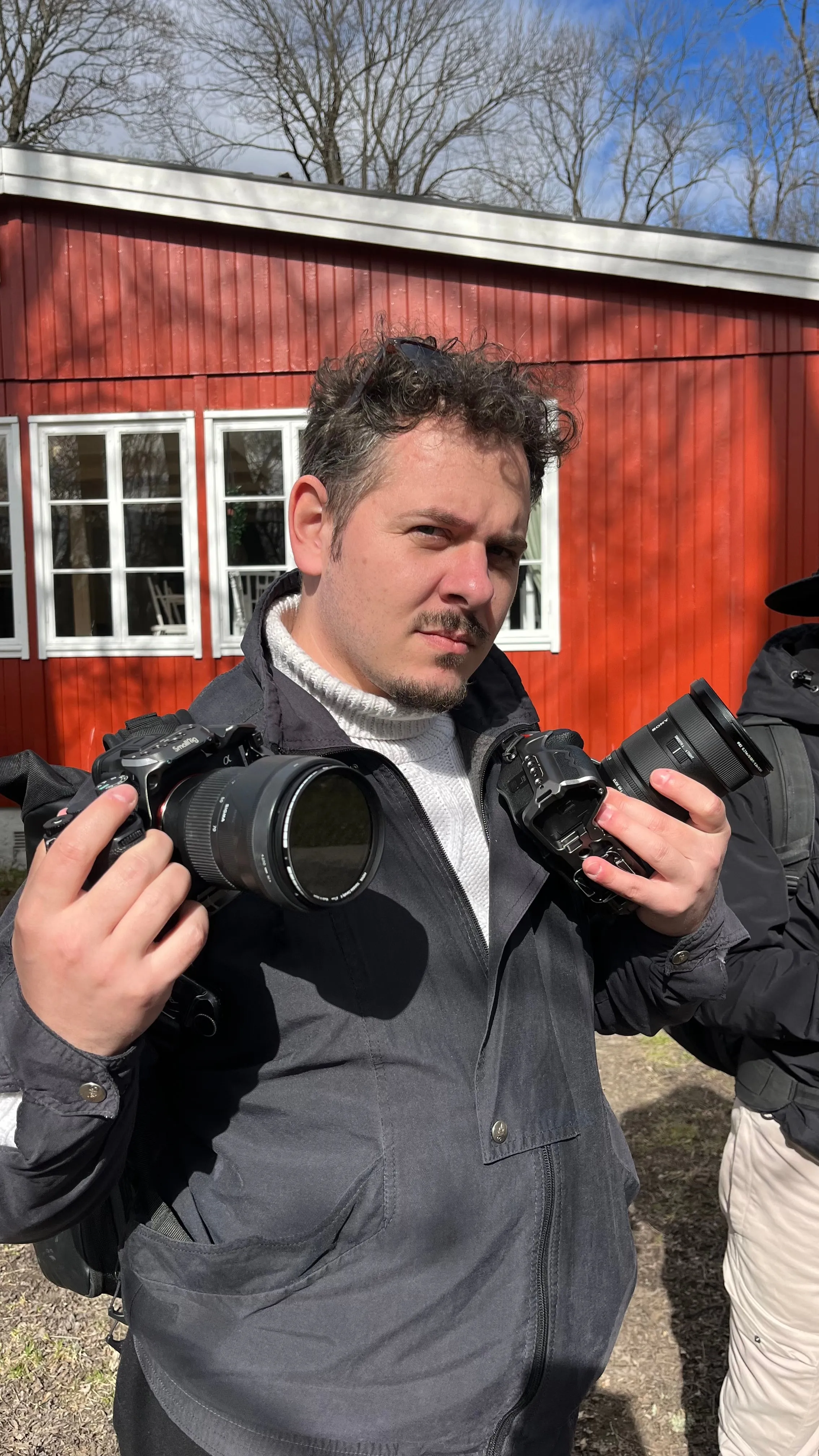 Un photographe tient un appareil dans chaque main devant une maison en bois rouge.