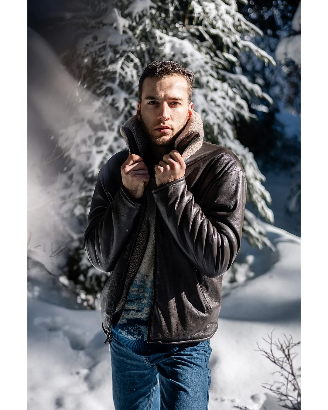 Un modèle en blouson de cuir remonte son col devant des sapins couverts de neige.