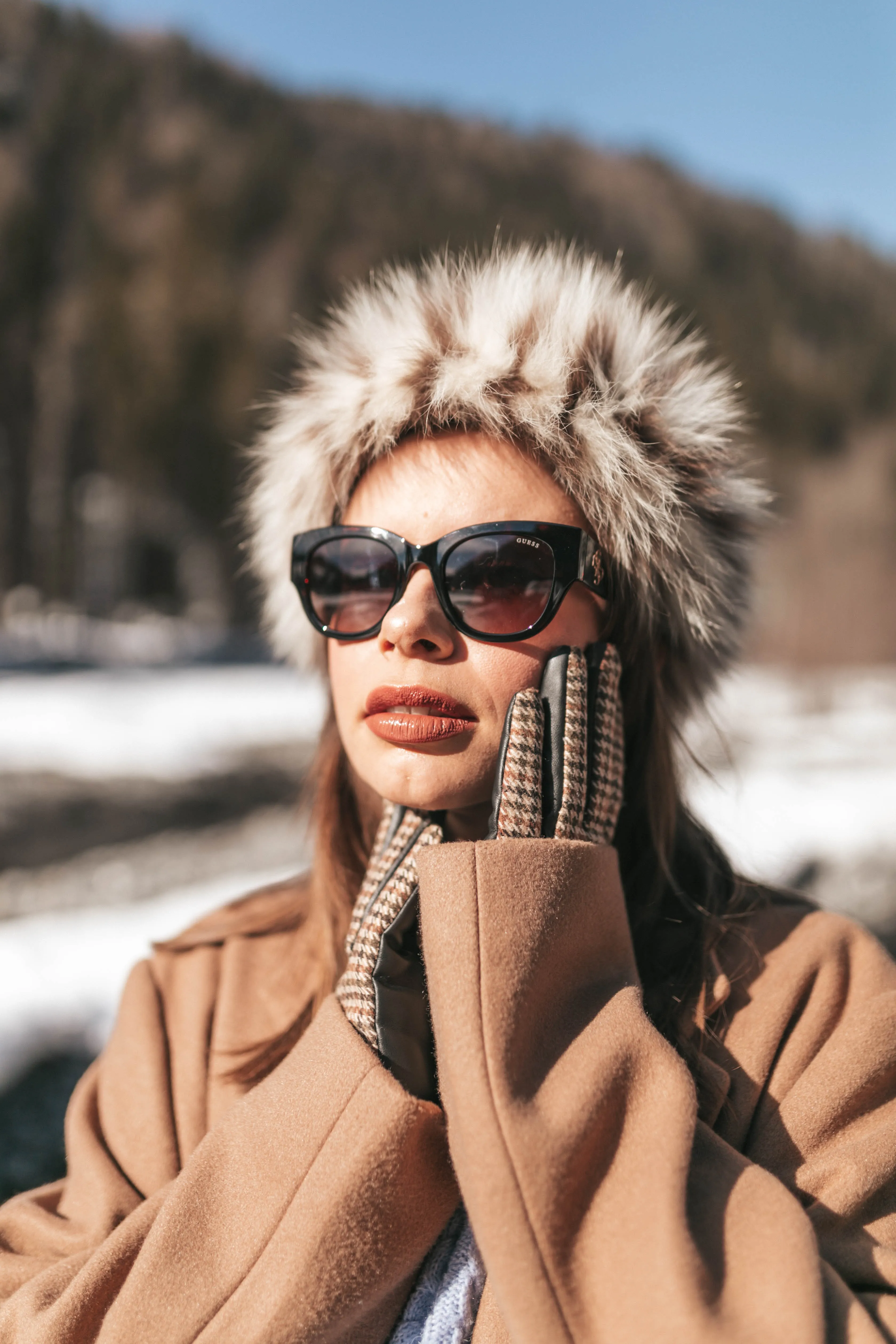 Une modèle en chapka de fourrure et lunettes de soleil, mains gantées contre le visage, dans la lumière d'hiver.