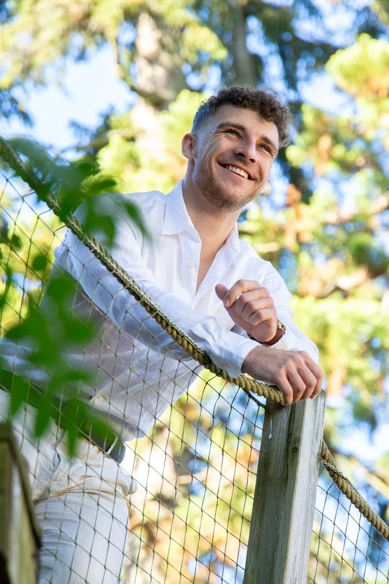 Un modèle souriant en chemise blanche, accoudé à une passerelle en bois au milieu des arbres.