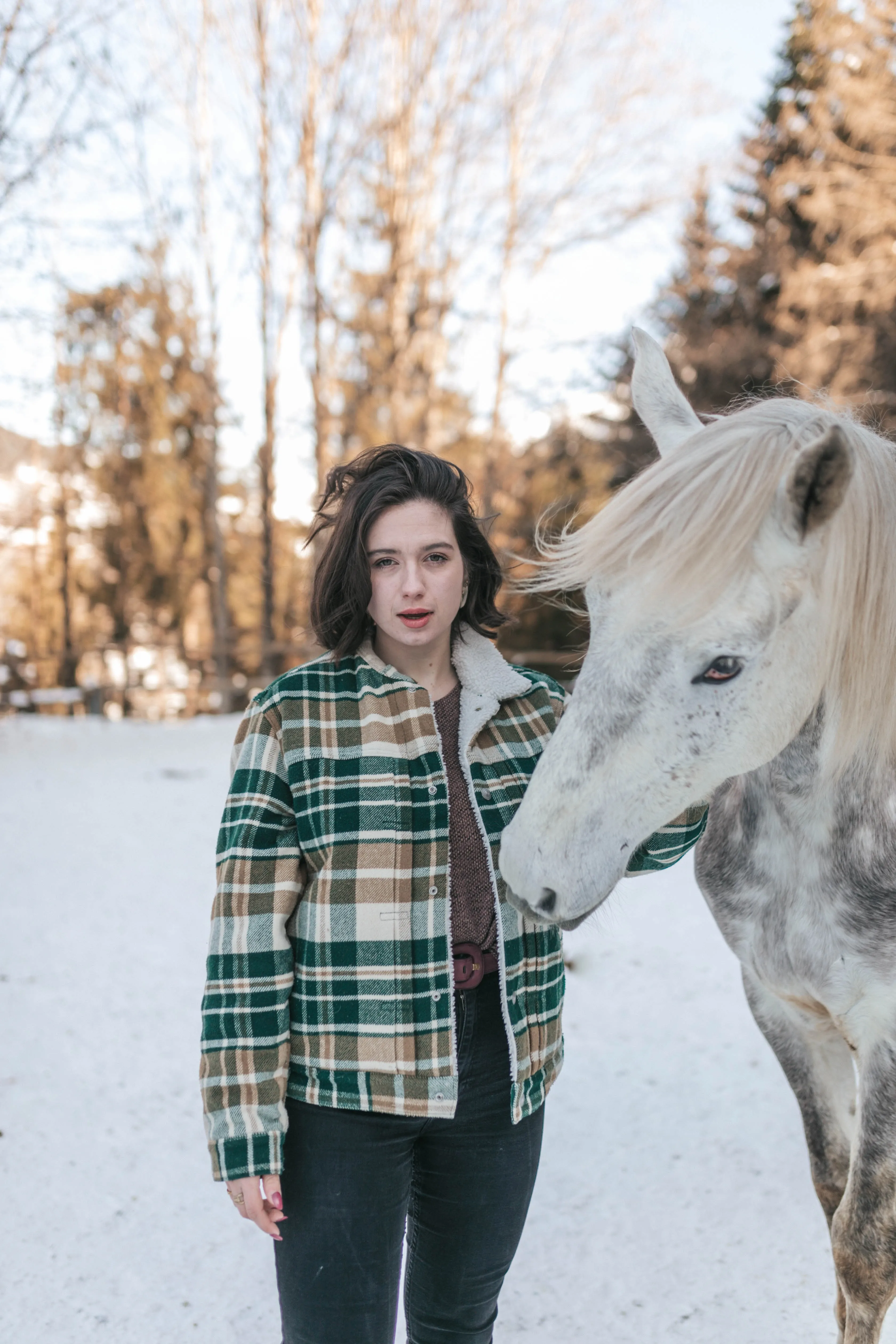 Une modèle en veste à carreaux pose à côté d'un cheval blanc, dans un paysage enneigé.