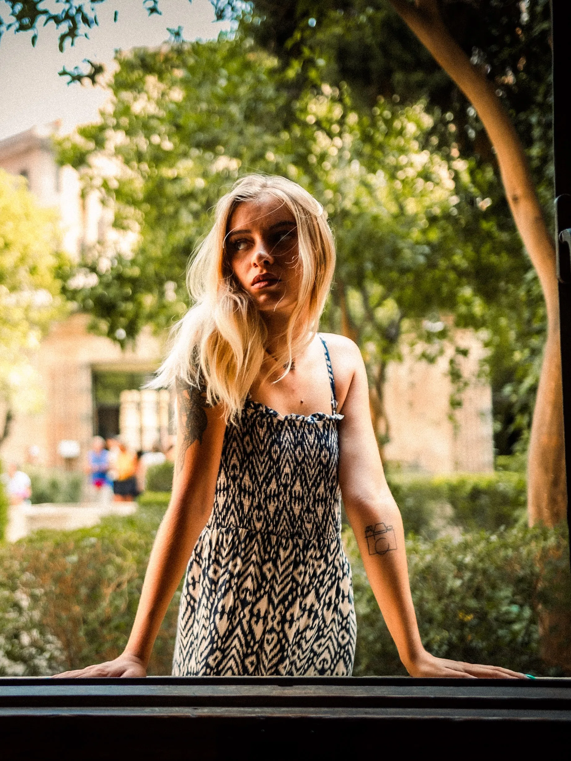 Une modèle blonde en robe à motifs s'appuie au rebord d'une fenêtre ouverte sur un jardin.