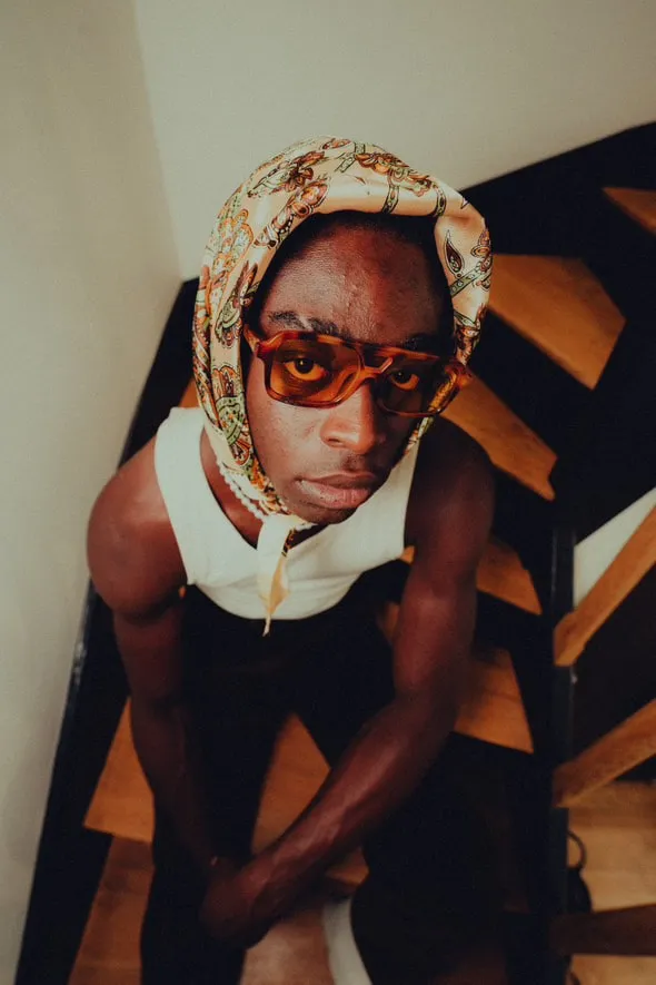 Un modèle en foulard fleuri et lunettes écaille, assis dans un escalier en bois.