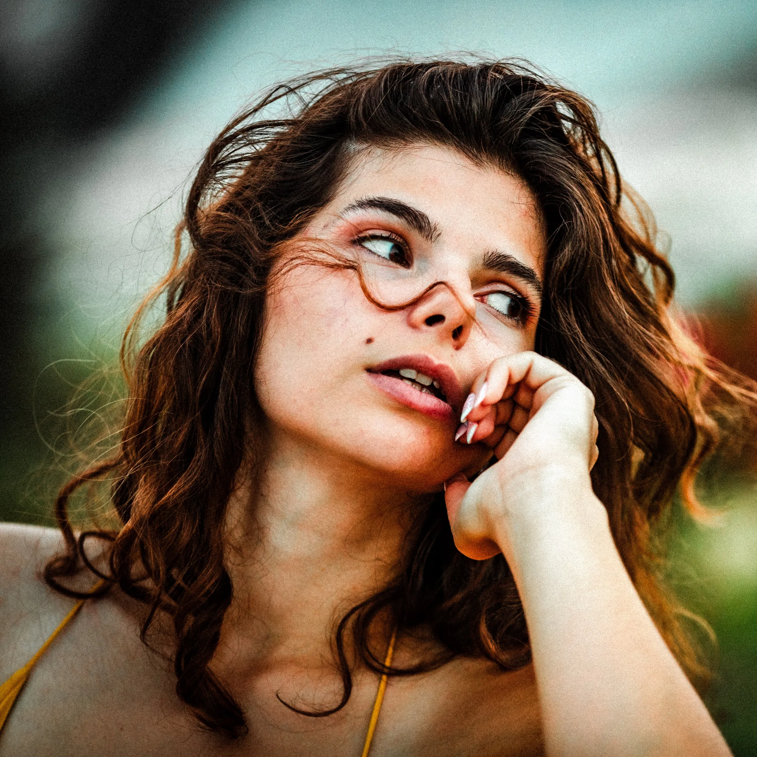 Gros plan d'une modèle aux cheveux bouclés et lunettes fines, la main contre la joue, devant des fleurs rouges floues.