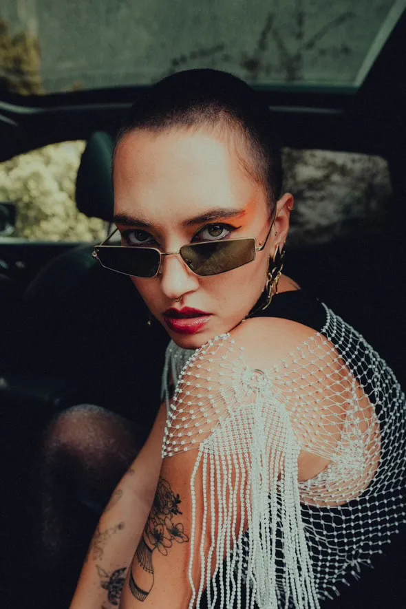 Une modèle aux cheveux rasés, lunettes fines et châle de perles, assise dans une voiture.