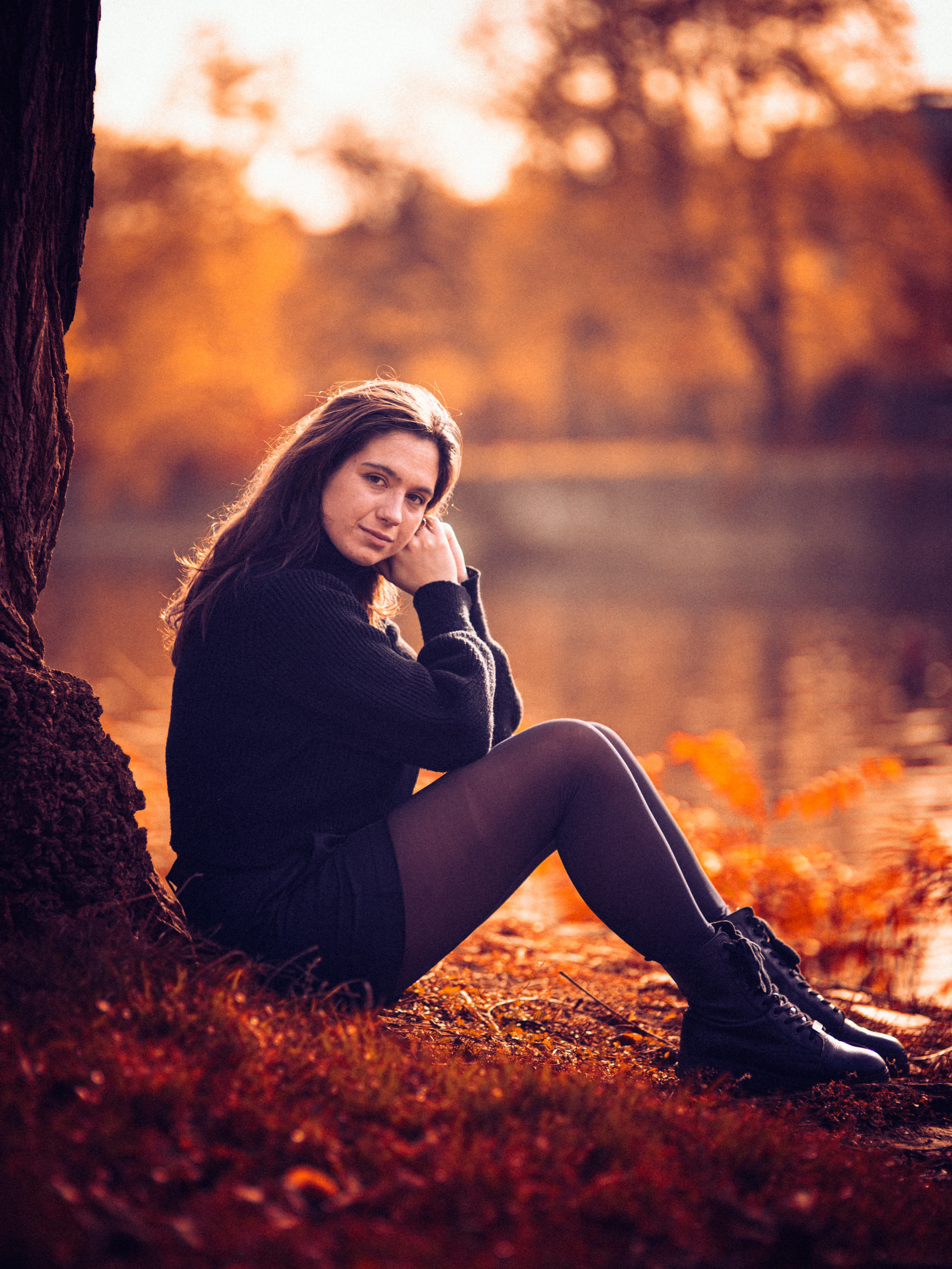 Une modèle en pull noir assise contre un arbre, au bord de l'eau, dans la lumière dorée d'automne.