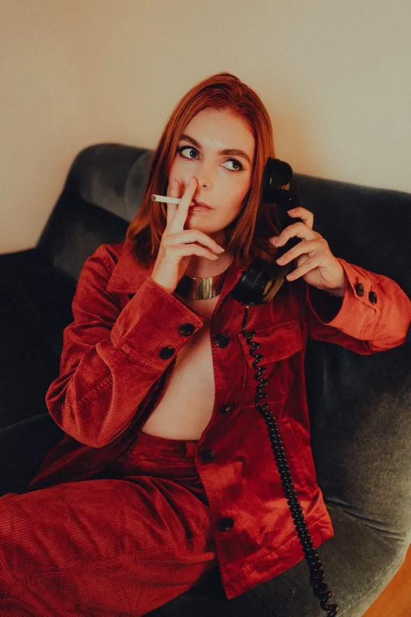 Une modèle rousse en ensemble rouge, cigarette et combiné de téléphone à la main, dans un fauteuil.