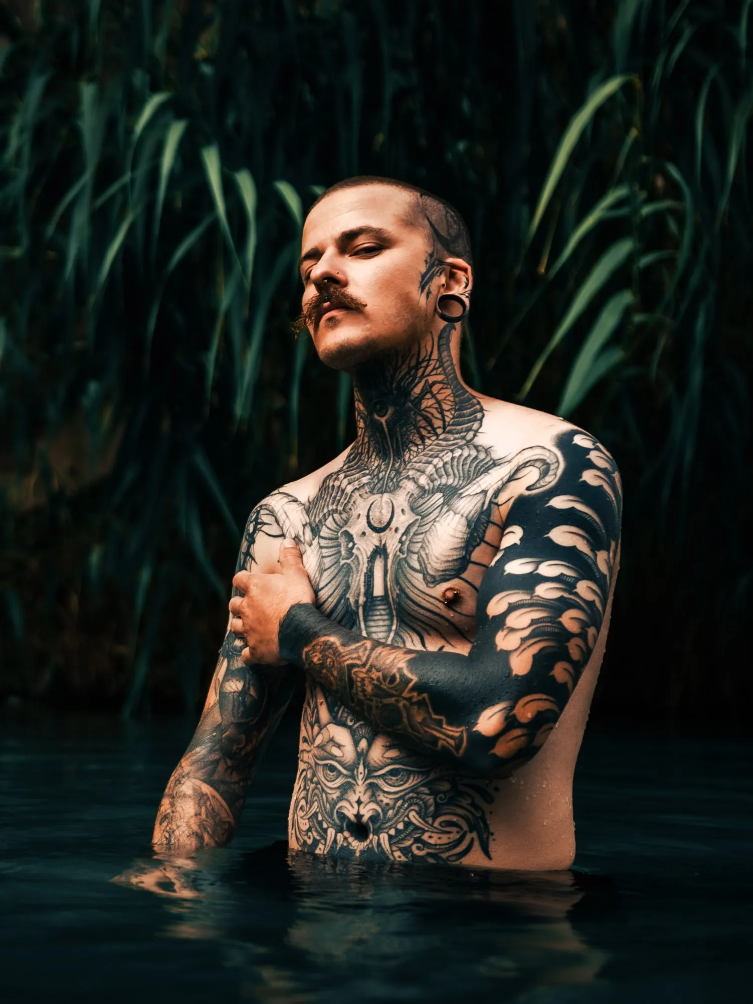 Un modèle au corps tatoué pose dans l'eau jusqu'à la taille, devant de hauts roseaux.