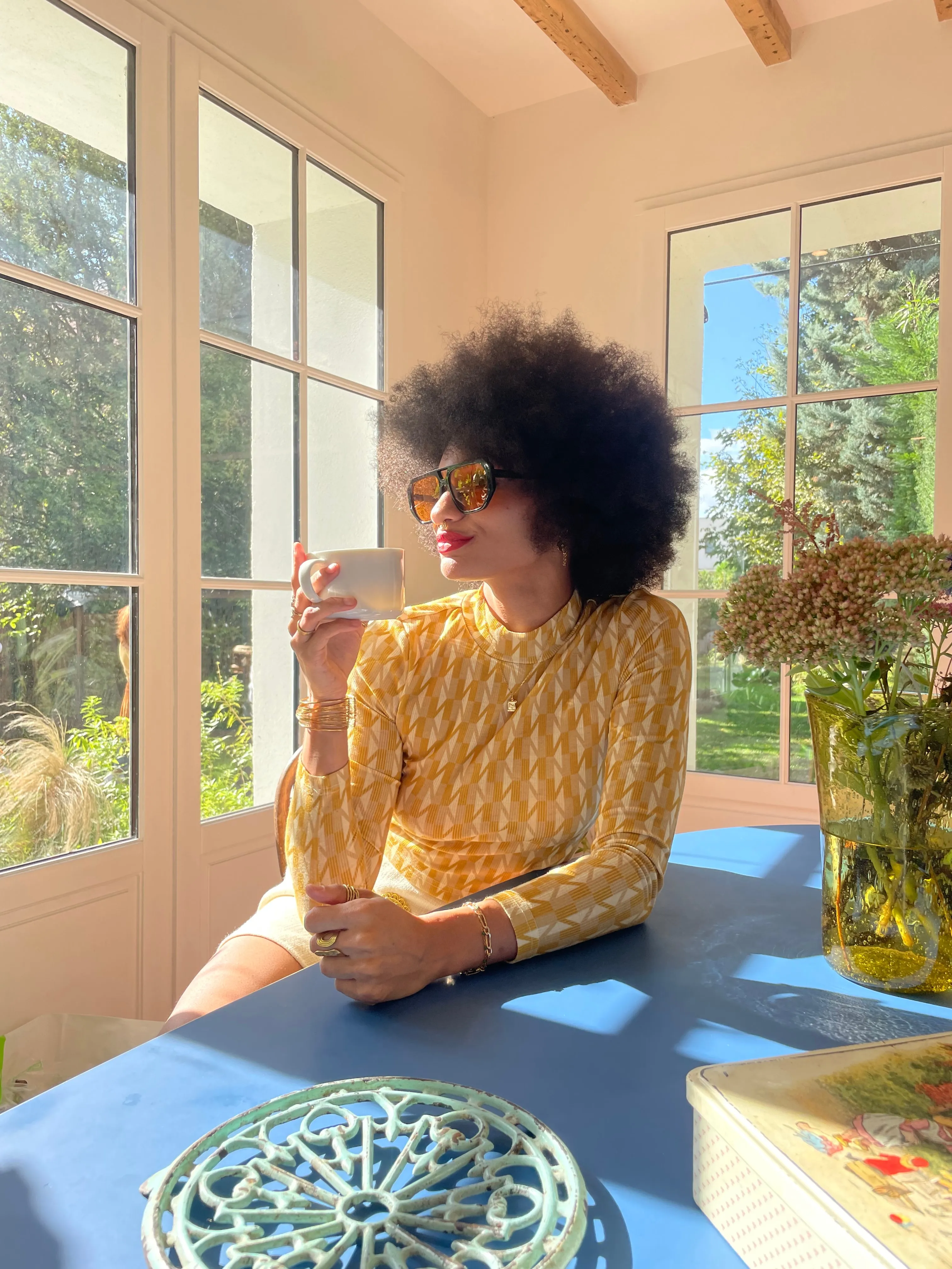 Une modèle aux cheveux afro, lunettes de soleil et chemisier jaune rétro, tasse à la main dans une véranda.