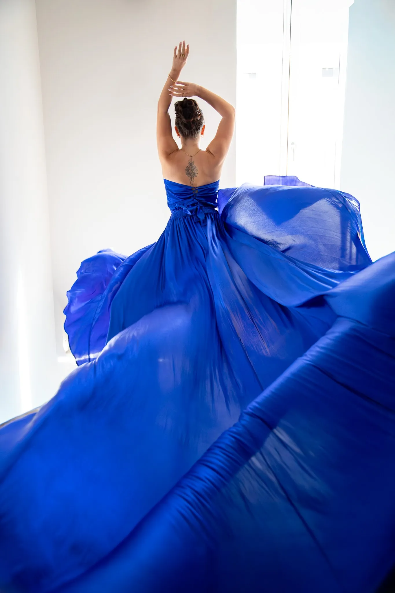 Une modèle de dos, bras levés, dans une robe bleu roi dont le tissu s'envole autour d'elle.