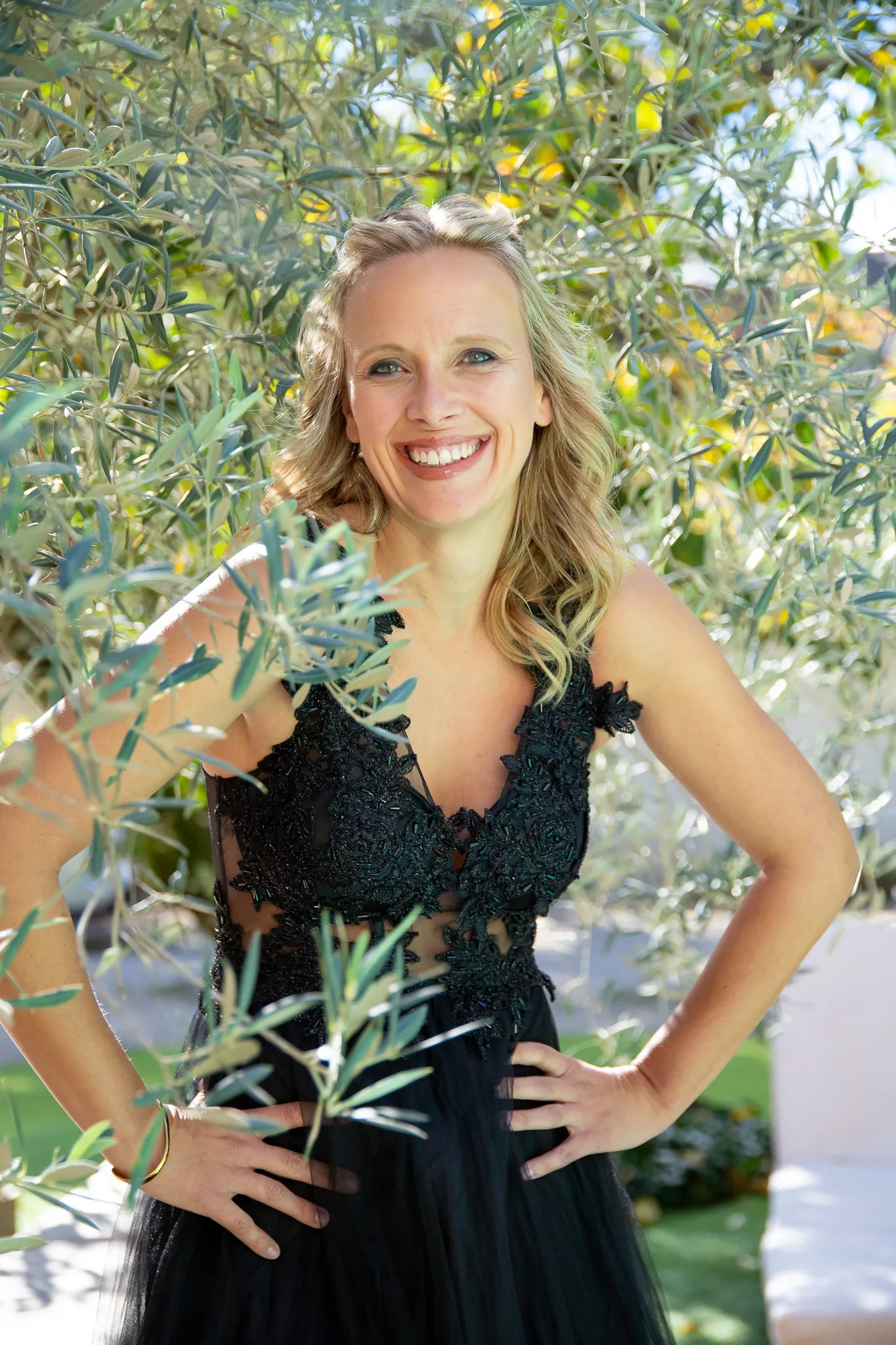 Une modèle souriante en robe noire en dentelle pose sous un olivier.