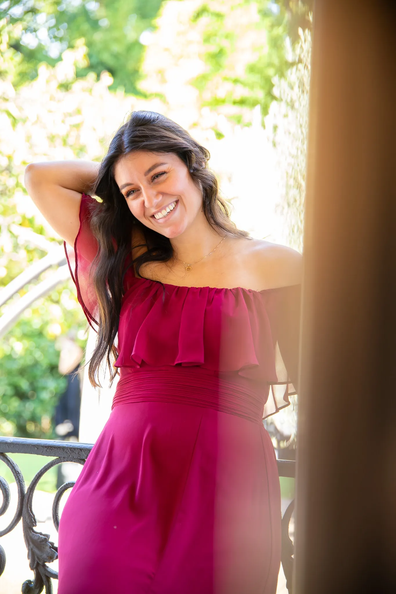 Une modèle en robe fuchsia à épaules dénudées rit, appuyée à un balcon en fer forgé.