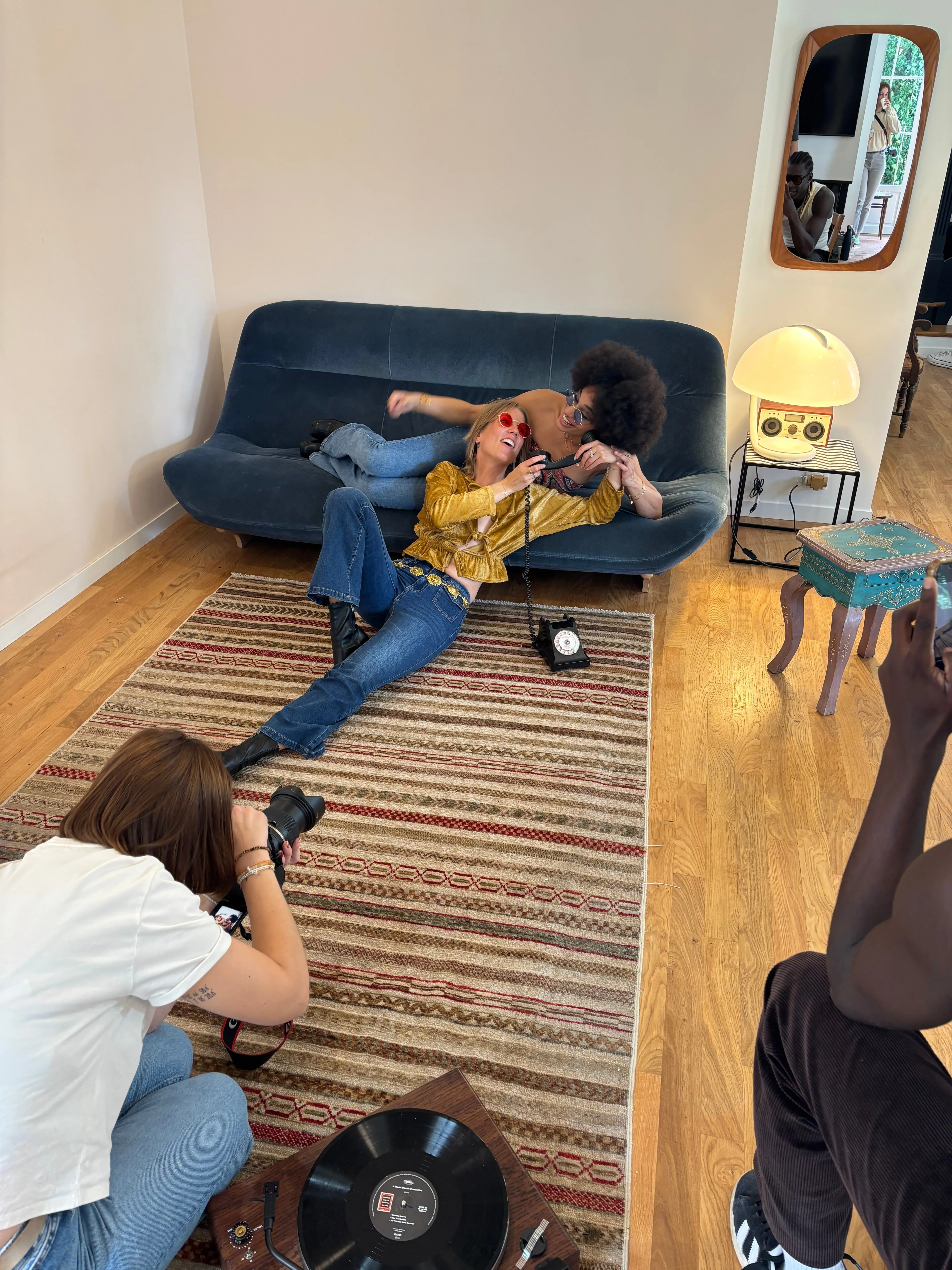 Une photographe au sol photographie une modèle allongée sur un canapé bleu, dans un décor rétro.