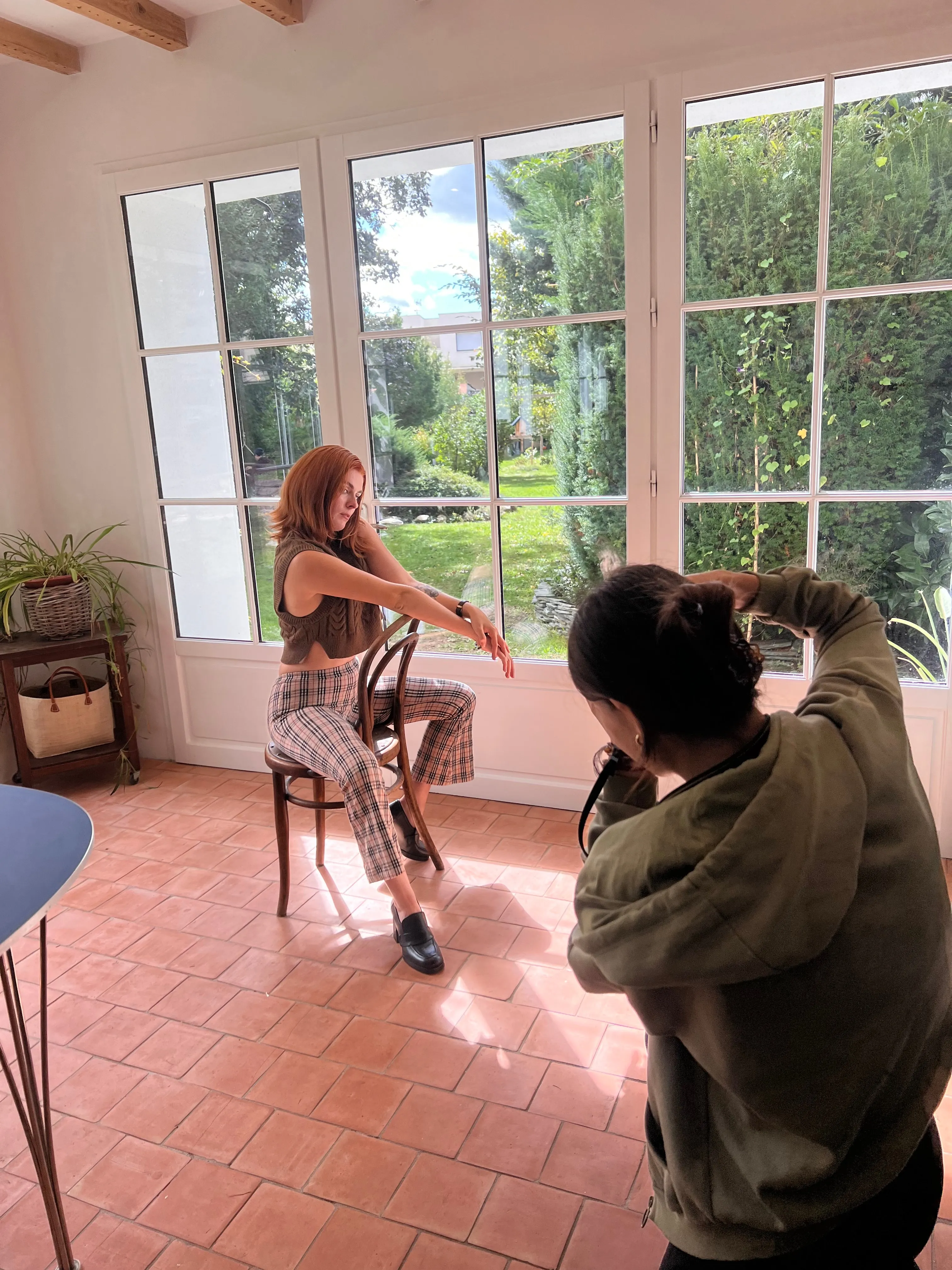 Une photographe cadre une modèle assise sur une chaise bistrot, devant les grandes fenêtres d'une véranda.