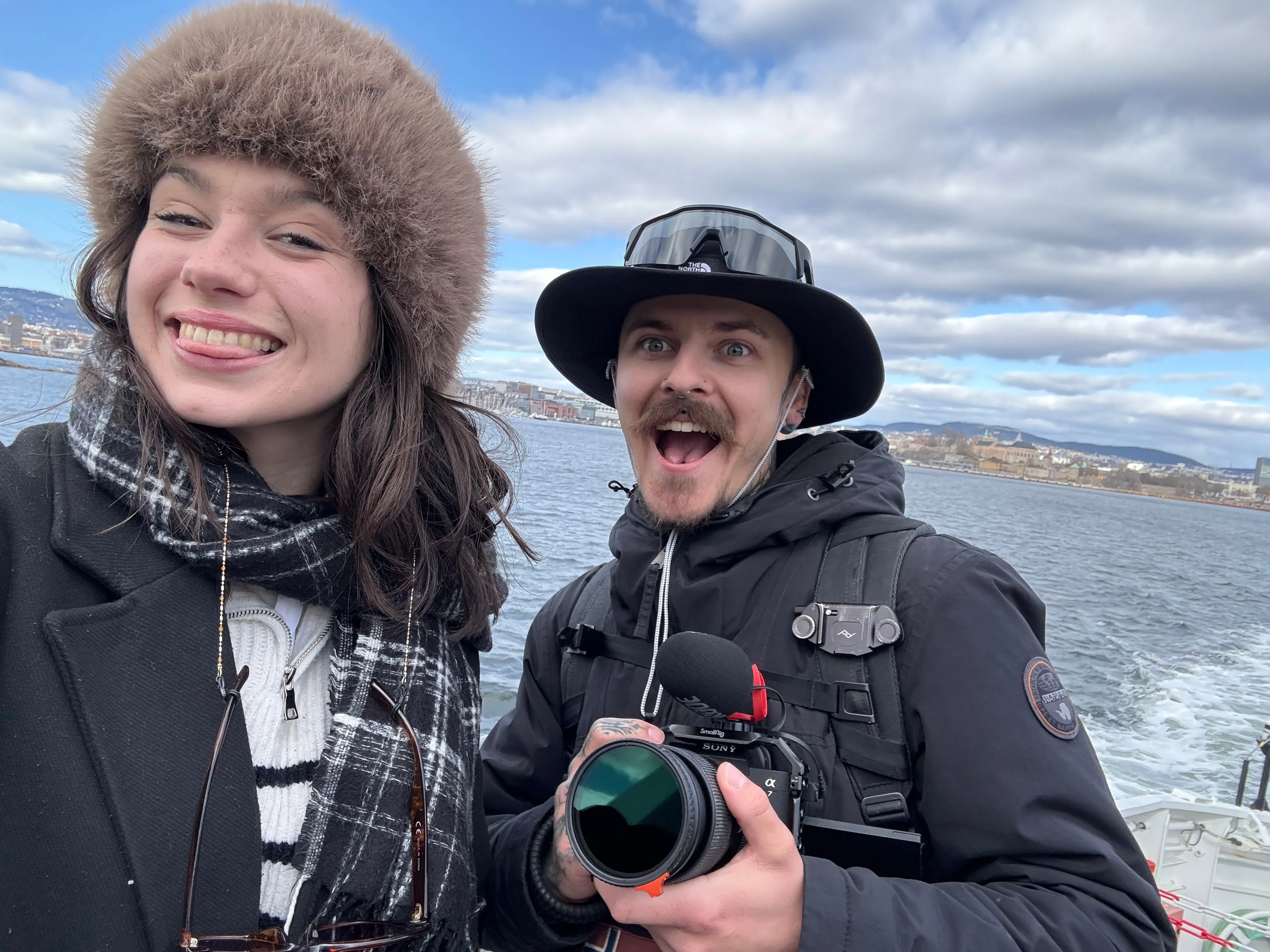 Deux participants rient sur le pont d'un bateau, appareil photo en main, avec la côte d'Oslo derrière eux.