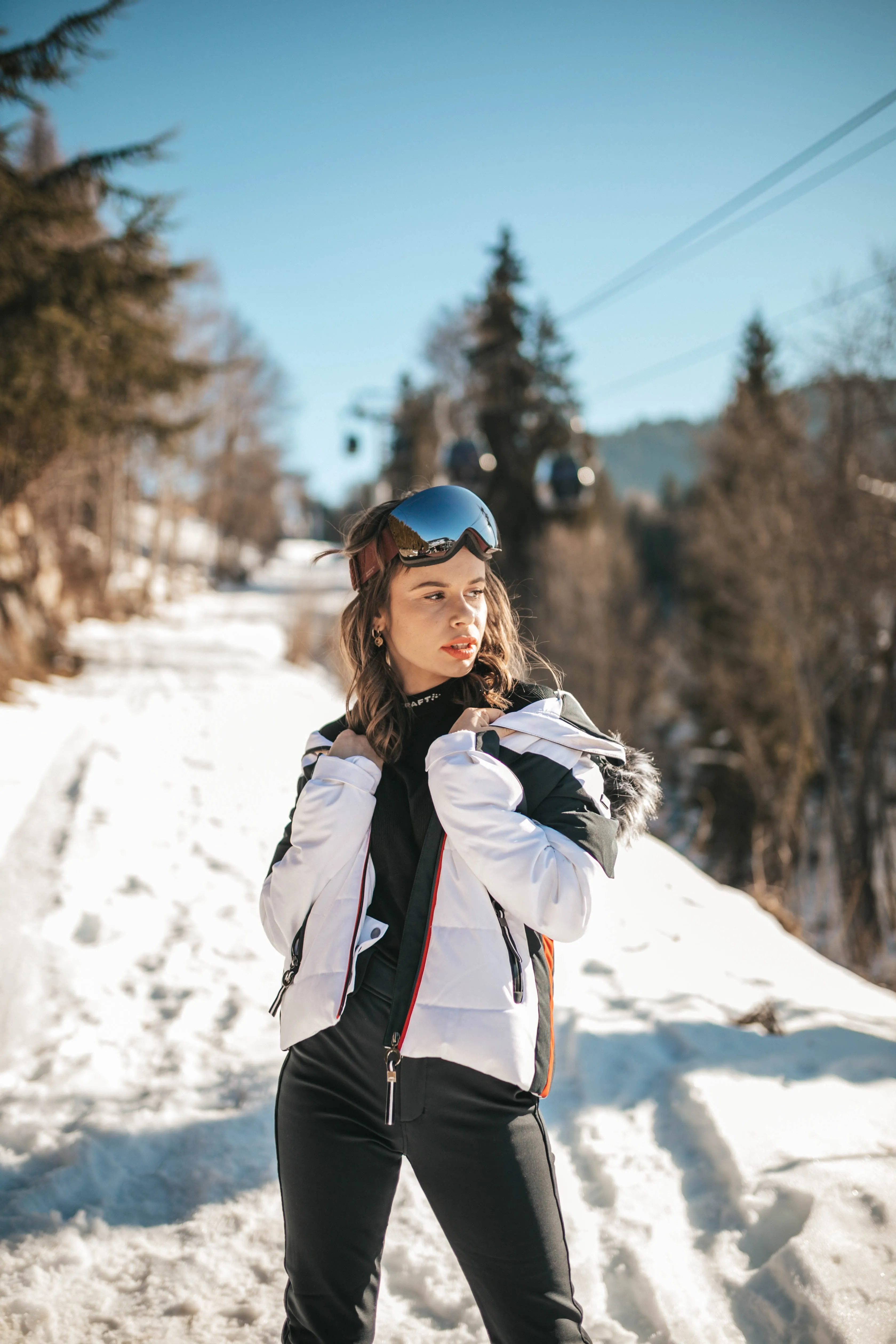 Une modèle en veste de ski blanche et masque sur le casque, sur un chemin enneigé bordé de sapins.